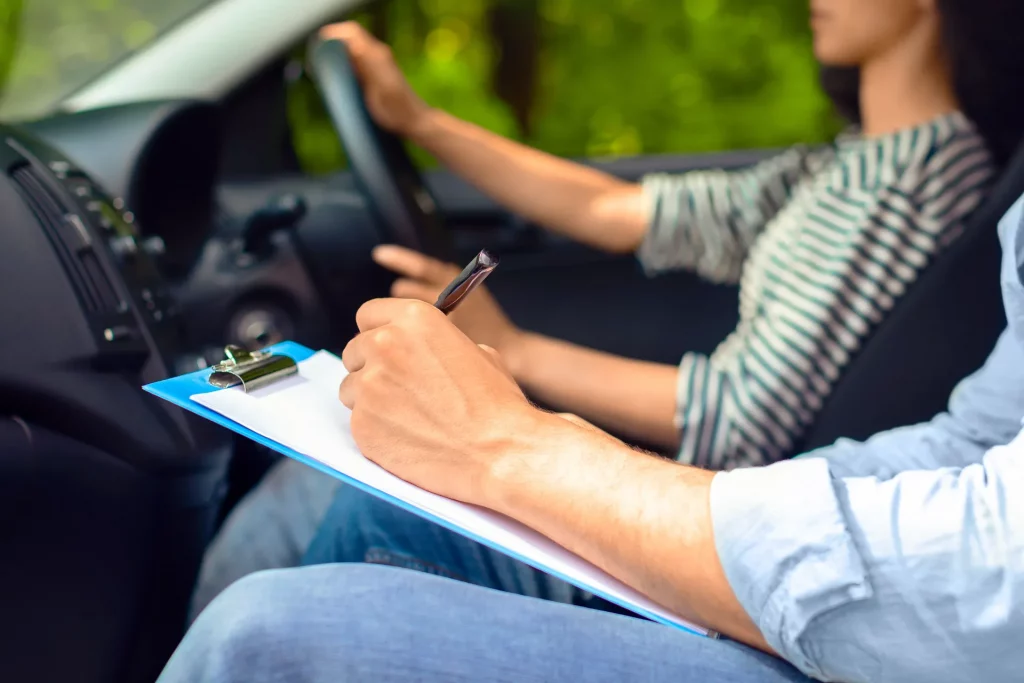 his image shows a driving instructor holding a clipboard while a learner drives an automatic car. It reflects a real driving lesson environment and supports the blog topic explaining how much are automatic driving lessons, what happens in a lesson
